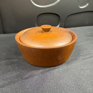 May include: A round, medium-brown wooden bowl with a matching circular lid. The lid features a small, centered, knob-style handle. The bowl has a smooth, tapered exterior surface showing a natural wood grain pattern throughout the entire piece.