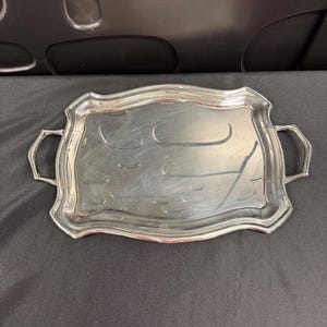 May include: A rectangular silver-toned serving tray with a polished, reflective surface and a scalloped edge. The tray features two integrated handles on the short sides and exhibits visible surface scratches across the center area.