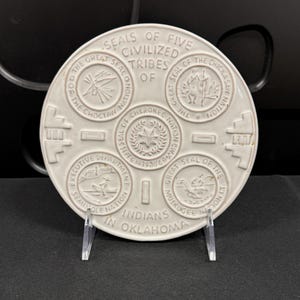 May include: A round, off-white ceramic commemorative plate featuring embossed text and imagery. The center displays the Great Seal of the Cherokee Nation, surrounded by the seals of the Choctaw, Chickasaw, Seminole, and Muskogee Nations.