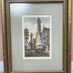Op de afbeelding: Een ingelijste ets met een Europees stadsplein met een hoge centrale toren, historische trapgevels en een fontein op de voorgrond. Het kunstwerk is gevat in een getextureerde goudkleurige en groene lijst met een lichtgrijze passe-partout.