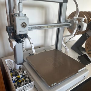 Puede incluir: Una impresora 3D Bambu Lab gris y blanca con una placa de construcción rectangular. La impresora tiene un sistema de pórtico y un contenedor de residuos blanco lleno de escombros de colores. Un rollo de filamento es visible en el fondo.