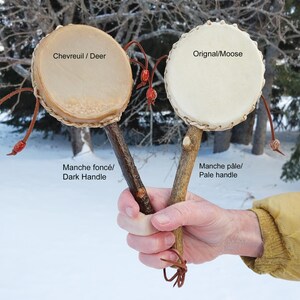 Könnte beinhalten: Zwei kleine, runde Trommeln mit hellen Trommelfellen und Holzgriffen. Eine Trommel ist mit "Chevreuil / Deer" und die andere mit "Orignal/Moose" beschriftet. Die Griffe sind in verschiedenen Brauntönen gehalten. Die Trommeln werden in einer Hand vor einem schneebedeckten Hintergrund gehalten.