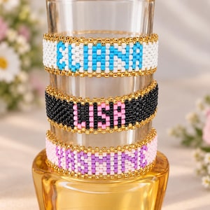 Könnte beinhalten: Drei Perlenarmbänder gestapelt auf einem Glaszylinder. Das obere Armband ist weiß mit dem Namen ELENA in leuchtendem Blau. Das mittlere Armband ist schwarz mit dem Namen LISA in Pink. Das untere Armband ist weiß mit dem Namen JASMINE in Lila.