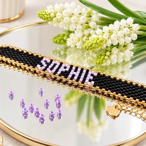 Könnte beinhalten: Ein Perlenarmband mit schwarzem Hintergrund und dem Namen SOPHIE, der aus hellvioletten Perlen besteht. Das Armband ist mit goldfarbenen Perlen eingefasst und hat einen kleinen herzförmigen Anhänger an einer goldfarbenen Metallkette.