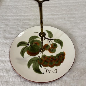 May include: A two-tiered serving stand with a white ceramic plate featuring a hand-painted fruit design in green, orange, and brown. The stand has a brass-colored handle and a brown rim. Ideal for displaying desserts or appetizers.