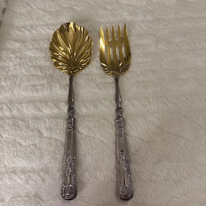 Peut inclure: Un ensemble de service composé d'une cuillère et d'une fourchette dorées. La cuillère a un design en forme de feuille et la fourchette a plusieurs dents. Les deux ont des manches argentés ornés de motifs décoratifs.