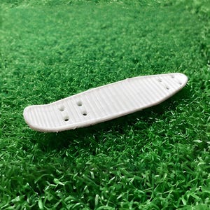 Könnte beinhalten: Ein weißes Skateboard-Deck mit strukturierter Oberfläche und vorgebohrten Löchern. Das Deck liegt auf einem Bett aus grünem Kunstrasen und erzeugt einen Kontrast zwischen den glatten Linien des Boards und dem strukturierten Hintergrund.