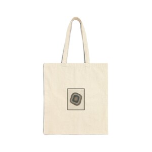 May include: A cream-coloured tote bag featuring a rectangular graphic of a layered, circular design in shades of grey, brown, and green, framed in a black square. The bag has a simple, minimalist design with long handles.