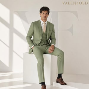 Puede incluir: Un traje de tres piezas verde salvia, que incluye chaqueta, chaleco y pantal&oacute;n, usado con una corbata melocot&oacute;n claro y una camisa blanca. El traje se combina con zapatos de vestir de cuero marr&oacute;n y calcetines negros. El logotipo de VALENFOLD es visible.