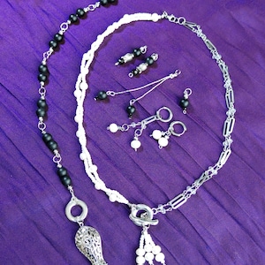 Puede incluir: Una colección de joyería hecha a mano, que incluye collares y pendientes, exhibida sobre tela morada. Los collares presentan cuentas negras y blancas, cadenas plateadas y colgantes decorativos. Los pendientes tienen varios diseños con cuentas y detalles plateados.