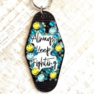 May include: Black keychain with the words "Always Keep Fighting" in white script. The keychain is decorated with yellow smiley faces and blue and teal rhinestones. A silver key ring is attached at the top.