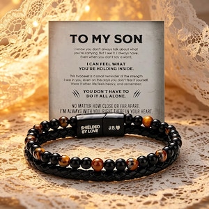 Peut inclure: Bracelet noir avec perles et pierres œil de tigre, et un bracelet en cuir tressé. Le fermoir magnétique porte les mots "SHIELDED BY LOVE" et "J.B.❤". Une carte avec le texte "TO MY SON" est visible en arrière-plan.