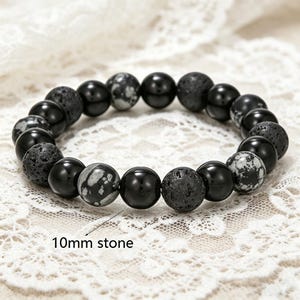 May include: A black beaded bracelet featuring 10mm stones. The bracelet includes a mix of polished black, speckled, and textured lava stone beads. The bracelet is displayed on a white lace surface.