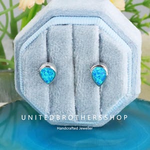 May include: A pair of silver stud earrings with teardrop-shaped, vibrant blue opal stones. The earrings are displayed in a light blue velvet jewelry box. The text "UNITED BROTHERS SHOP" and "Handcrafted Jeweller" are visible.