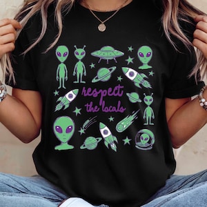 Könnte beinhalten: Schwarzes T-Shirt mit einem Design aus grünen Aliens, Raumschiffen, Planeten und Sternen. Der Schriftzug "respect the locals" ist in lila gehalten. Eine verspielte, Weltraum-Themen Grafik.