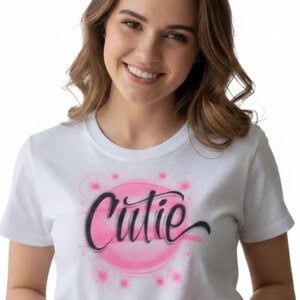 May include: White t-shirt with the word "Cutie" in a black script font, set within a pink circle with pink starburst accents. The shirt is short-sleeved and has a crew neck.
