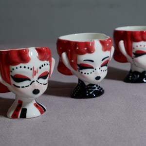 May include: Three ceramic egg cups shaped like stylised heads with red hair, white skin, and black facial features. Each cup features a unique painted design on the base, including black and red patterns, and a smooth, glossy finish throughout.