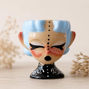 May include: A ceramic egg cup shaped like a stylized head with light blue hair, a tan face, and closed eyes. The figure features a vertical dotted line down the center of the face, pink blush on the cheeks, and a black base with white wavy lines.