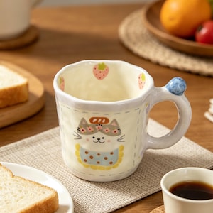 May include: A cream-colored ceramic mug with a cat design. The mug features a cat face with a floral headband and a blue polka-dot bib. Strawberries decorate the rim. The handle has a blue berry accent. The mug sits on a neutral-colored placemat.
