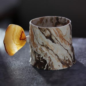 May include: A handmade ceramic mug with a unique agate handle. The mug features a marbled pattern in shades of brown and white. The agate handle is a translucent, golden-yellow color, adding a natural and artistic touch to the design.