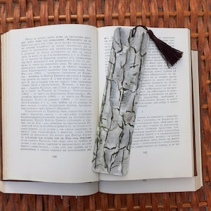 May include: A handmade bookmark featuring a tree bark design. The rectangular bookmark has a dark tassel. It's placed on a book, bringing a natural element to reading.