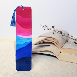 May include: A rectangular bookmark featuring a gradient of colors transitioning from deep red and magenta at the top to light blue, medium blue, and dark navy blue at the bottom. A bright blue tassel is attached to the top hole of the bookmark.
