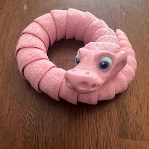 Può includere: Un giocattolo a forma di drago rosa articolato con una superficie strutturata e dettagliata. Il drago è arrotolato in un cerchio, con la testa sollevata e con occhi blu. Il giocattolo è fatto di plastica e poggia su una superficie di legno.