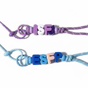 Könnte beinhalten: Zwei Schlüsselbänder mit Perlen, die Persönlichkeitstyp-Akronyme zeigen. Das obere Schlüsselband zeigt die Buchstaben ISFJ in weißen, lila und lavendelfarbenen Perlen mit einer lila Kordel. Das untere Schlüsselband hat ESFP in blauen und pfirsichfarbenen Perlen mit einer hellblauen Kordel.