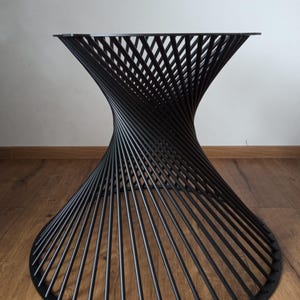 May include: Black metal table base with a unique, spiral design. The base features a circular bottom, a flared, twisted midsection, and a flat top, creating a modern, sculptural look. The table base is suitable for a variety of table tops.