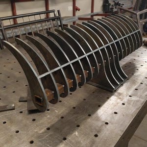 May include: A large, welded metal sculpture with a curved, rib-like design. The piece is made of thick, unfinished steel and sits on a metal work table. The sculpture's form suggests an abstract, industrial aesthetic.