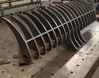 May include: A large, welded metal sculpture with a curved, rib-like design. The piece is made of thick, unfinished steel and sits on a metal work table. The sculpture's form suggests an abstract, industrial aesthetic.