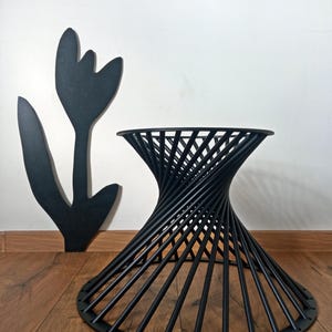 May include: Black metal side table with a distinctive spiral design. The table features a circular top and a flared base. A black tulip-shaped wall decoration is to the left. The table is on a wooden floor.