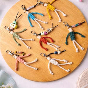 Può includere: Sette portachiavi a forma di bambola in macramè con chiusure a moschettone argentate, caratterizzati da corpi in corda colorata, teste in perline di legno e perline con lettere che compongono parole come LOVE, MARIA e BRIDE su legno.