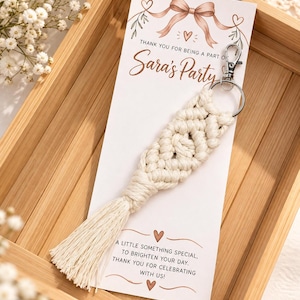 Peut inclure: Un porte-clés en macramé couleur crème avec un mousqueton et un anneau en métal argenté, fixé sur une carte rectangulaire blanche avec une illustration de nœud rose et le texte "Thank you for being a part of Sara's Party" et "A little something special, to brighten your day. Thank you for celebrating with us!"