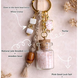 May include: A gold keychain with a turtle charm, a small glass bottle filled with pink salt, and a natural jute braided cord with a wooden bead. The keychain also features a glow-in-the-dark bead initial and white crystal.