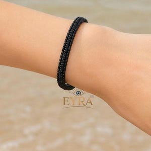 May include: A black bracelet featuring a woven or braided texture with a consistent, uniform pattern throughout the band. The accessory has a slim, rounded profile and a matte finish, positioned on a wrist with the text EYRA UK visible below.