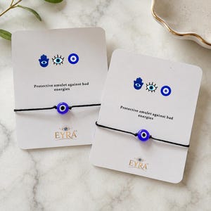 May include: Two black string bracelets with a blue glass evil eye bead, each mounted on a white display card featuring a hamsa, eye icon, and the text Protective amulet against bad energies and the brand name EYRA with an eye logo.