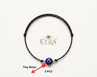 May include: A black cord bracelet featuring a single blue, white, and black glass evil eye bead centered between two small silver-toned metal beads. The text EYRA UK is printed above the bead, and the text Tiny Nazar 2 PCS is below.