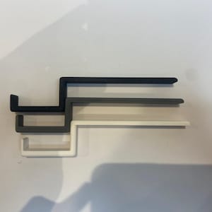 May include: Three L-shaped wall shelves in black, grey, and white are stacked. The shelves are made of a matte material and are mounted on a white wall. The design is minimalist and modern, suitable for various rooms.