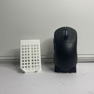 Pode incluir: Um rato de computador preto com uma roda de rolagem branca e botões laterais, ao lado de um suporte de telefone retangular branco com vários orifícios circulares. O rato está sobre uma base preta.