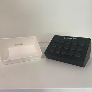 May include: A black Stream Deck control console with fifteen square buttons and a white rectangular tray with a small horizontal cutout on the back wall. The console features the Stream Deck logo printed in white on the top center edge.
