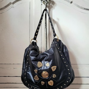 May include: Black leather hobo bag with gold-colored embellishments. The bag features a rounded shape, studded trim, and decorative gold accents in various shapes. The bag hangs from a metal hook.
