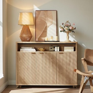 May include: A light wood cabinet with vertical ribbed doors and a top shelf displaying decor. A woven lamp with a beige shade sits on the cabinet. A framed print, candles, and a floral arrangement are also visible.