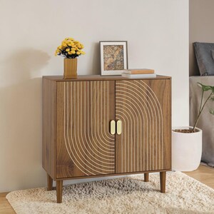 May include: A wooden cabinet with two doors and gold-colored handles. The cabinet has a brown finish with a white, curved design on the doors. A vase of yellow flowers and a framed picture sit on top of the cabinet.