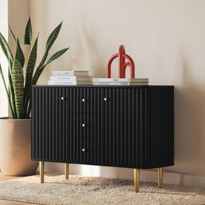 May include: A black, three-drawer cabinet with vertical ribbed detailing. The cabinet has gold-colored knobs and legs. A red sculpture and several books sit on top of the cabinet. A potted snake plant is to the left.