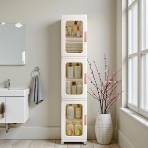 May include: A tall, white bathroom storage cabinet with three sections, each with a clear door. The cabinet contains toiletries, towels, and bottles. The cabinet has rose gold handles and is in a bathroom setting.