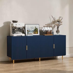 May include: A navy blue sideboard with four doors and gold-colored legs and handles. A white espresso machine, framed landscape art, and a cup rack with white cups are on top. Dried flowers in a glass vase complete the decor.