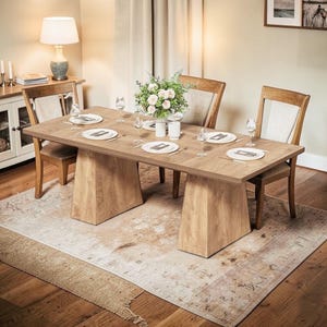 May include: A light brown wooden dining table with a rectangular top and two trapezoidal supports. The table is set with plates, silverware, and wine glasses for six people. A floral arrangement sits in the center. The table is on a patterned rug.