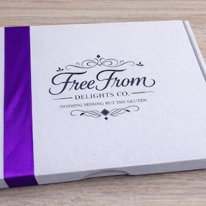 Puede incluir: Una caja blanca con una cinta morada en el lado izquierdo. La caja tiene las palabras "Free From Delights Co." y "Nothing missing but the gluten" impresas en la parte superior en escritura negra.