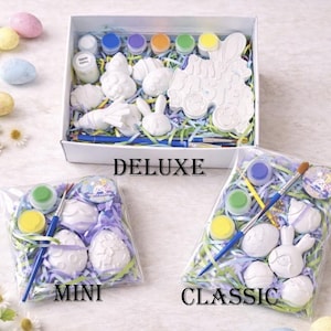 May include: Easter paint kits in three sizes: Deluxe, Mini, and Classic. The Deluxe kit includes ceramic shapes, paint pots, and brushes, all in a white box. The Mini and Classic kits are packaged in clear bags. The word "DELUXE", "MINI", and "CLASSIC" are printed on the image.